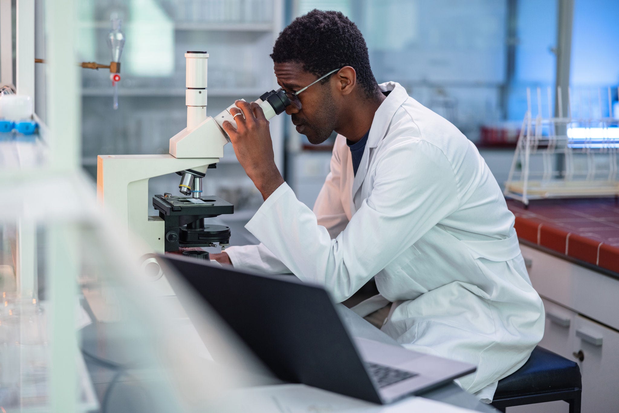 Portrait of African-American male scientist wearing a lab coat, looking under a microscope, analyzing a sample during his work in a laboratory.