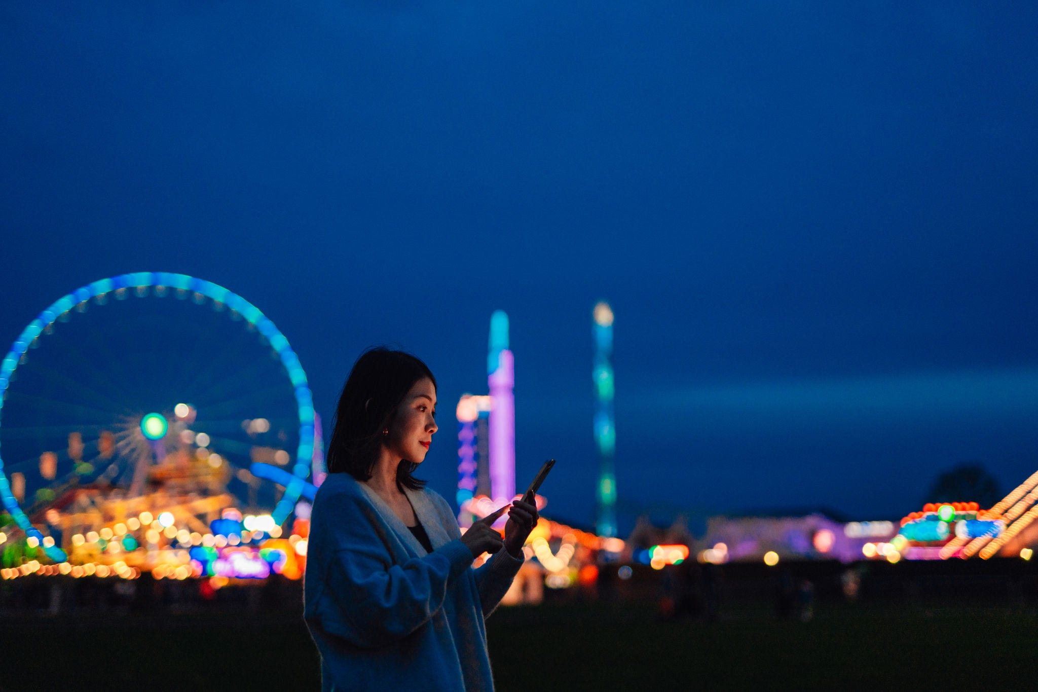 Side view of beautiful young Asian woman looking aways while using smartphone in carnival at night with illuminated night in the background. Mobile banking. Business on the go.