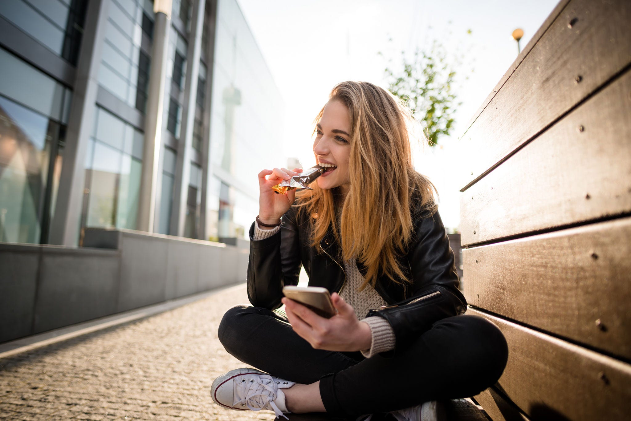 Wide angle photograph of teenager eating chocolate on phone outddoor in street sitting on bench